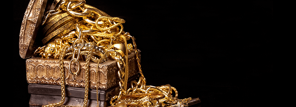 gold bracelets, rings, necklaces piled up and tumbling out of a small wooden jewelry box in front of a black background