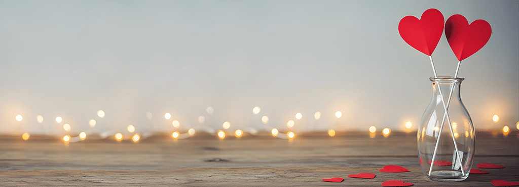 glase vase with 2 red hearts mounted on the top of sticks sticking out of it on a wooden table top with fairy lights in the background